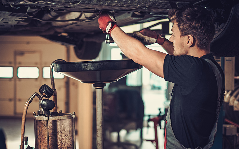 A mechanic in gloves working under a car, draining oil into a pan.