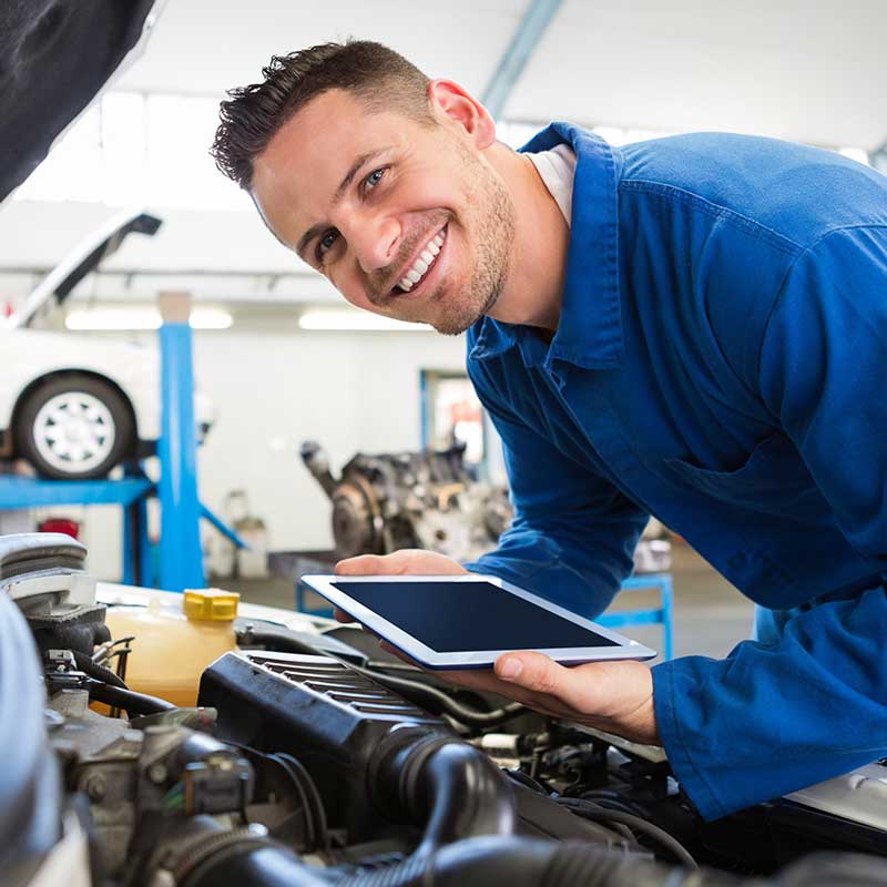A smiling Acura mechanic in a blue uniform holds a tablet while working on a car engine.